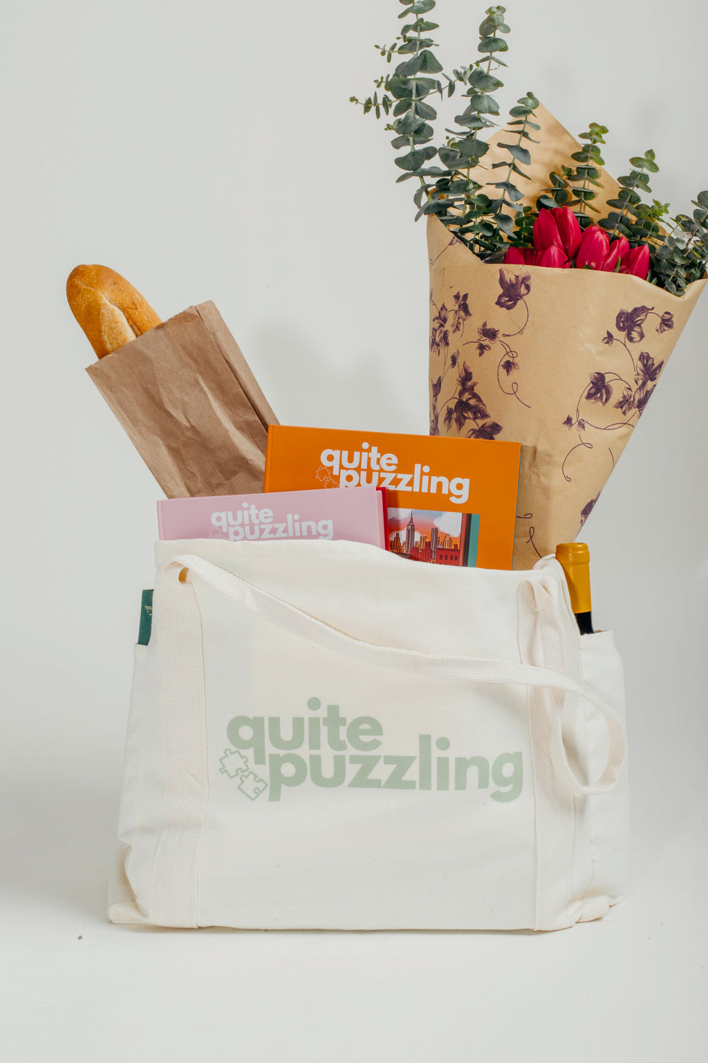 White tote bag with 'quite puzzling' branding, containing a loaf of bread, magazine, and flowers.