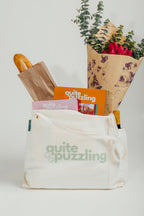White tote bag with 'quite puzzling' branding, containing a loaf of bread, magazine, and flowers.