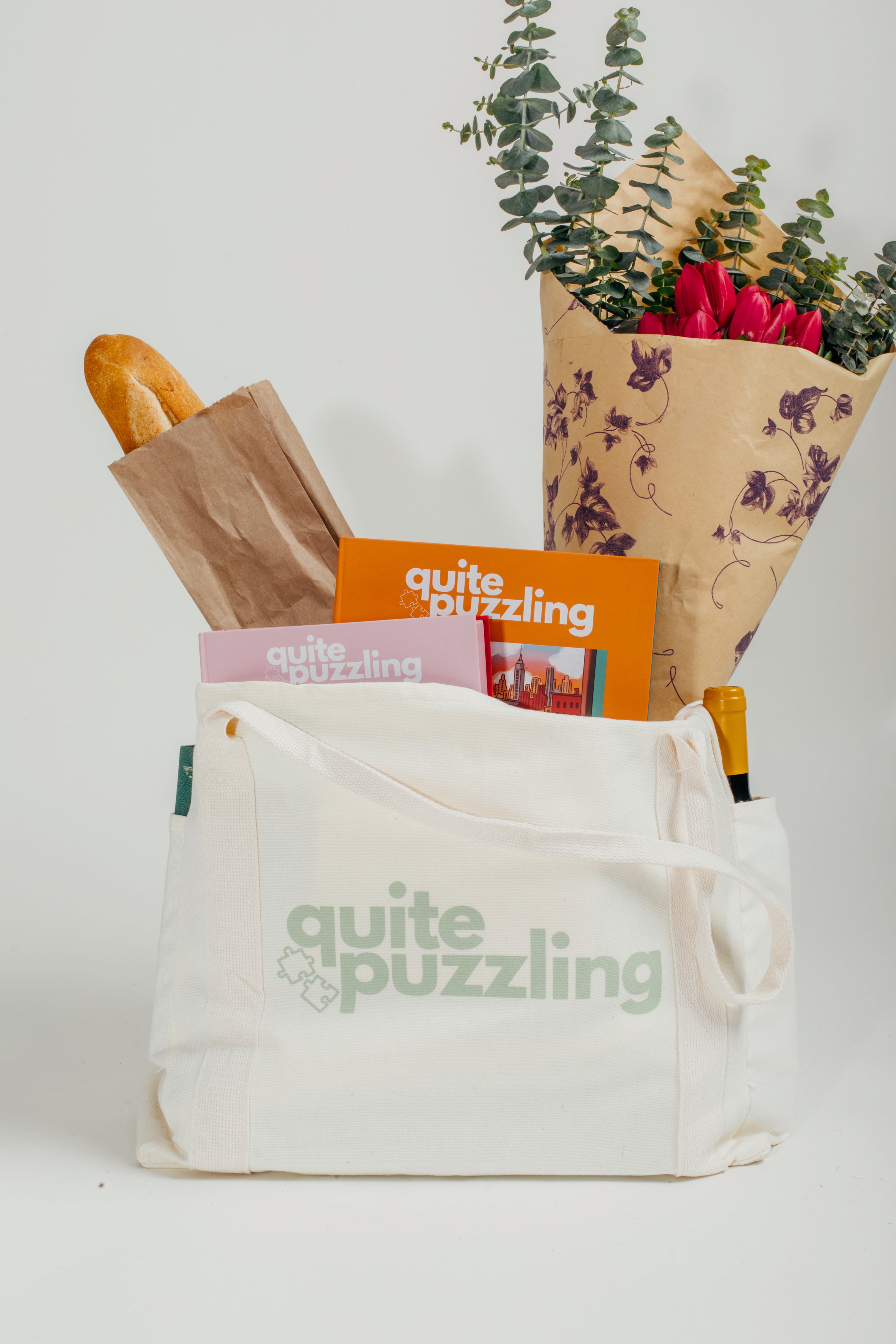 White tote bag with 'quite puzzling' branding, containing a loaf of bread, magazine, and flowers.